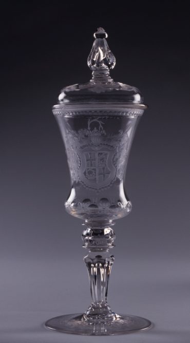 Das Bild zeigt den Deckelpokal. Es handelt sich um einen gläsernen Pokal, auf dem das Wappen des Landkomturs Karl Heinrich von Hornstein eingraviert ist. Der Pokal schließt mit einem ebenfalls aus Glas bestehenden Deckel ab.