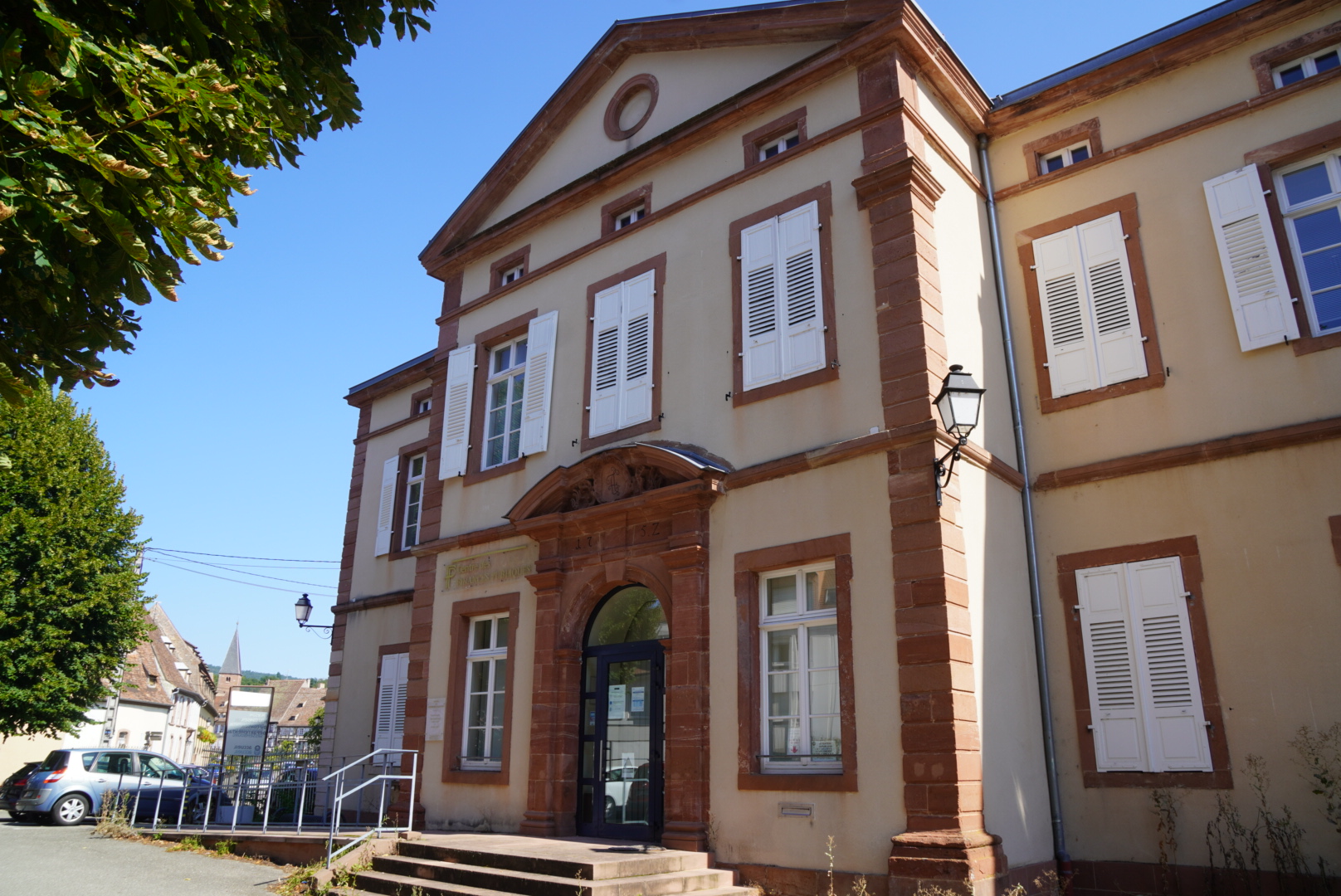 Das Bild zeigt ein Gebäude in Weißenburg, in dem sich heute verschiedene Behörden befinden. Das Gebäude hat viele hohe Fenster mit weißen Fensterläden. Sowohl am Eingang, der die Form eines Torbogens, hat als auch an weiteren Stellen des Hauses ist rötlicher Sandstein angebracht.