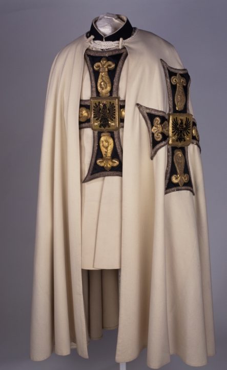 Das Bild zeigt das Ornat des Hochmeisters Erzherzog von Österreich. Es handelt sich dabei um ein cremfarbenes Gewand, auf dem an der Seite und vorne auf der Brust Deutschordenskreuze aufgenäht sind.
