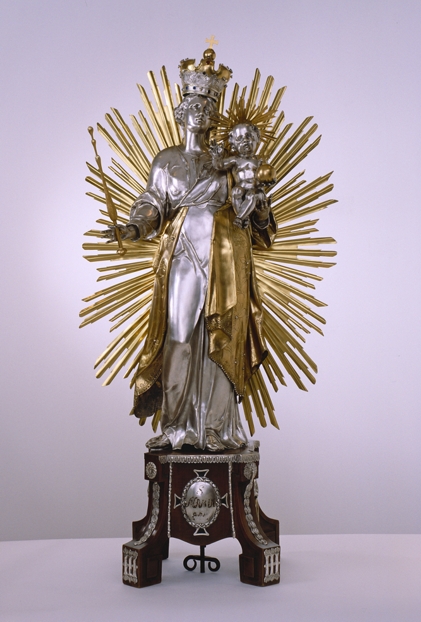 Das Bild zeigt eine Silberfigur der Mutter Gottes mit Jesuskind auf dem linken Arm, Lilienstab in der rechten Hand und einer Krone auf dem Kopf. Hinter ihr befindet sich ein vergoldeter Strahlenkranz. Die Figur der Mutter Gottes steht auf einem hölzernen Sockel, auf dem silberne Verzierungen angebracht sind.