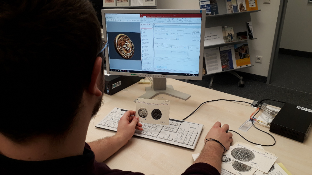 Auf dem Bild sieht man einen Mitarbeiter, der eine Karteikarte aus einer Siegelsammlung in der Hand hält und auf seinem Computer die Informationen von der Karteikarte in die Datenbank einträgt.
