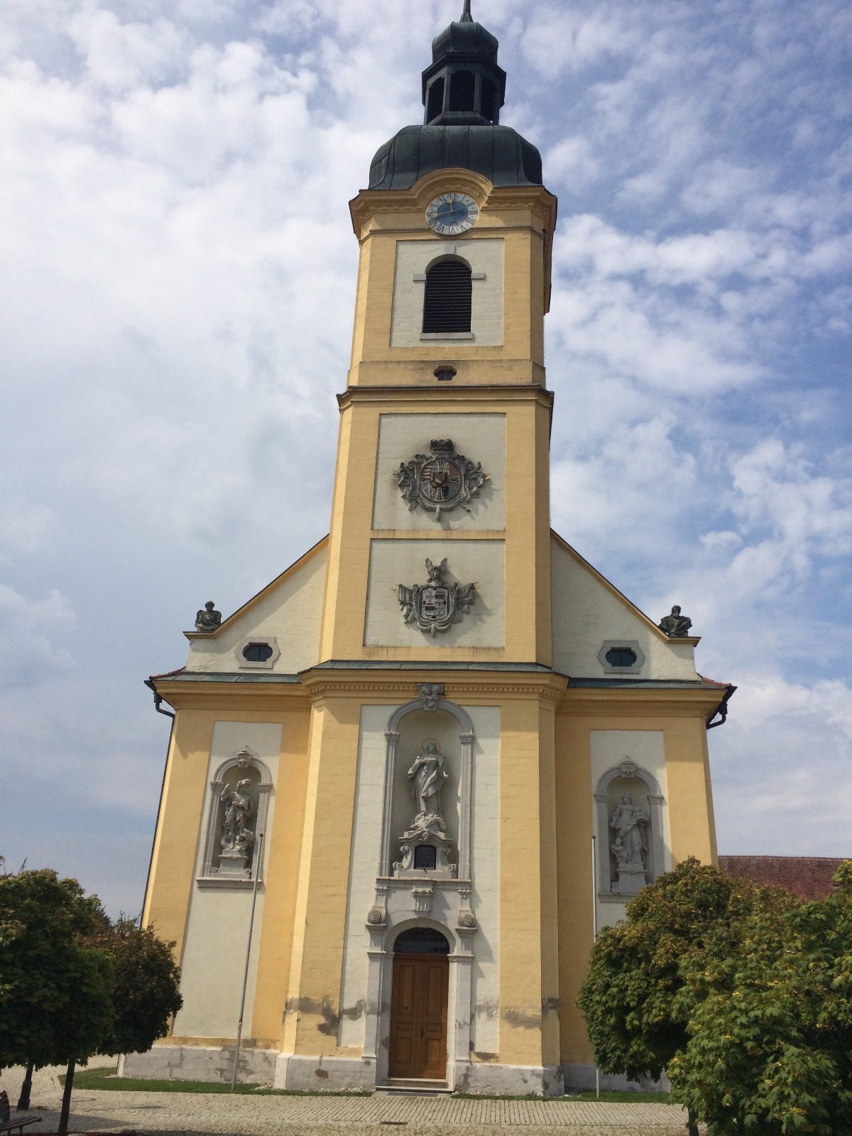 Auf dem Bild sieht man die St. Augustinus Kirche in Stopfenheim, die in gelb und weiß gehalten ist und eine graue Turmspitze besitzt.