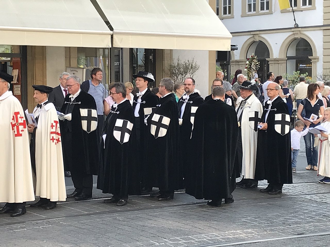 Das Bild zeigt Familiare des Deutschen Ordens sowie Templer in Ordenstracht, die bei einer Prozession in der Stadt teilnehmen. Um sie herum befinden sich weitere Personen.