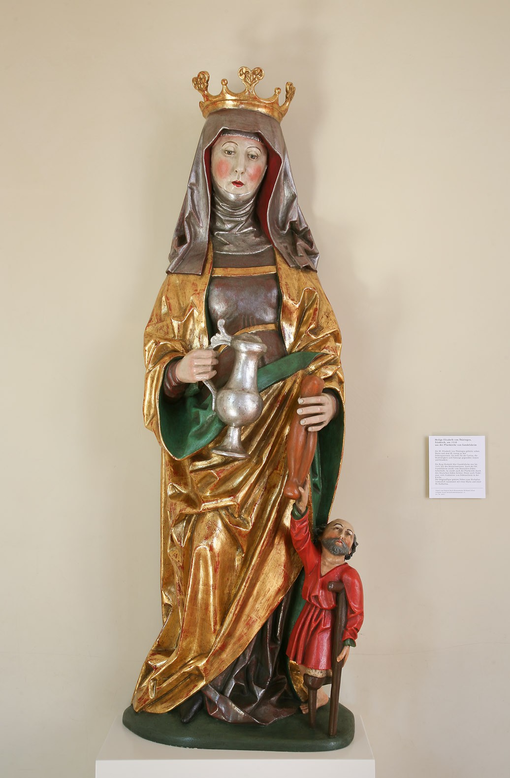 Das Bild zeigt eine Figur der Heiligen Elisabeth von Thüringen. Sie trägt ein Tuch auf dem Kopf, auf dem außerdem eine Krone sitzt, und hält einen Krug in der rechten Hand. An ihrer linken Seite befindet sich die Figur eines kleinen Mannes, der aufgrund eines fehlenden Beins auf Krücken gestützt ist.