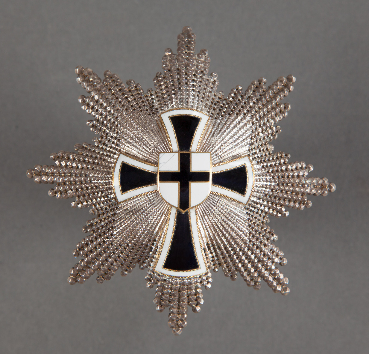 Das Bild zeigt das große Verdienstkreuz des Deutschen Ordens. Zentral ist darauf ein Deutschordenskreuz zu sehen auf dem ein Deutschordenswappen angebracht ist. Das Kreuz ist auf einem aus Strahlen zusammengesetzten Stern befestigt.