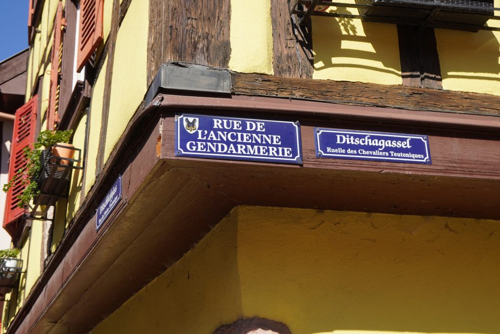 Das Bild zeigt ein Straßenschild auf Elsässisch und Französisch auf dem man die Geschichte des Deutschen Ordens in Kaysersberg erkennen kann.
