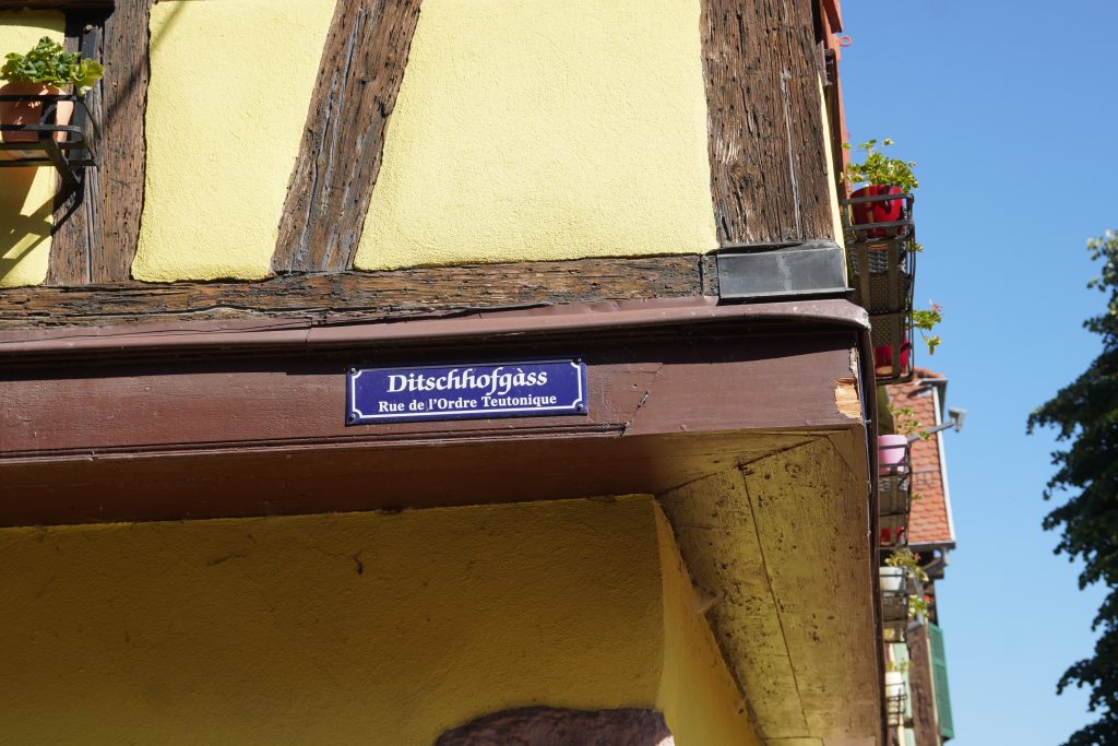 Das Bild zeigt ein Straßenschild auf Elsässisch und Französisch auf dem man die Geschichte des Deutschen Ordens in Kaysersberg erkennen kann.